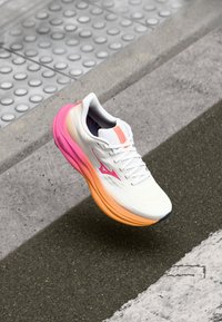 White running shoe with pink and orange accents, mesh upper, cushioned sole, and textured details. Positioned mid-air above a pavement.