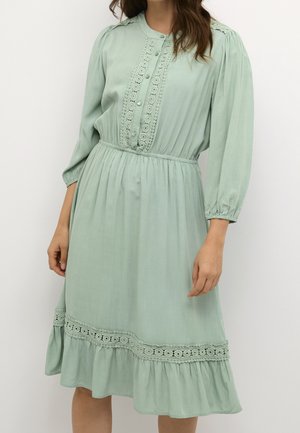 Kobieta w jasnooliwkowej sukience midi z koronkowym wykończeniem, guzikami na przodzie, długimi rękawami oraz marszczonym stanem i dołem.