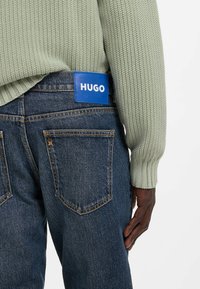 Zielony, dziergany sweter i ciemnoniebieskie dżinsy z widoczną niebieską metką HUGO na tylnej części pasa. Teksturowana tkanina i zarysowany krój.