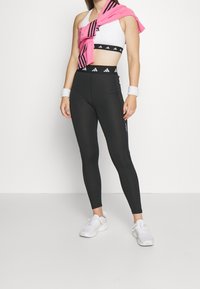 Leggings atletici neri con vita alta, abbinati a un reggiseno sportivo bianco con una canotta a strisce nere e rosa legata sulla spalla.