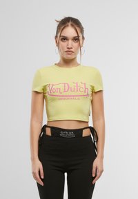 Gul avkortet t-skjorte med korte ermer som har en rosa "Von Dutch Originals" logo over forsiden. Svart bukse med utskjæringsdetaljer.