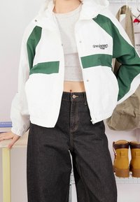 Biała kurtka wiatrówka z ciemnozielonymi panelami, zamkiem błyskawicznym i kapturem. Nosi się ją na szarym topie typu crop, w połączeniu z ciemnymi spodniami z wysokim stanem.