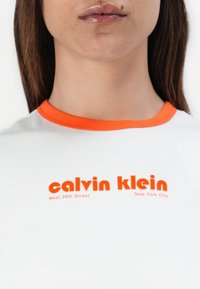 White t-shirt with an orange crew neck and orange text featuring "calvin klein" and "West 39th Street, New York City." Smooth fabric texture.