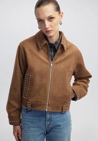 Veste bomber en suede marron ornée de studs argentés le long du col et des poignets, fermeture éclair à l'avant, accompagnée d'une chemise à carreaux bleus.