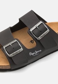 Chancla negra con dos correas ajustables, hebillas metálicas, plantilla de corcho y logo en relieve. Cuenta con una suela texturizada para mayor agarre.