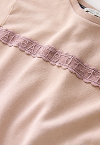 Šviesiai rožinis megztinis, puoštas tekstūruota, banguota nėrinių juosta su išsiuvinėtomis raidėmis ant krūtinės. Minkšta medžiaga su lygiu paviršiumi.