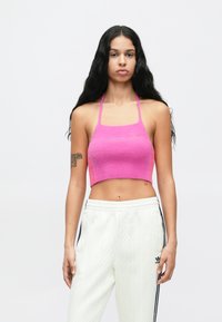 Jovem mulher com cabelo longo e preto, vestindo um top curto rosa com alças e calças de malha brancas da Adidas com riscas pretas, em pé contra um fundo branco.