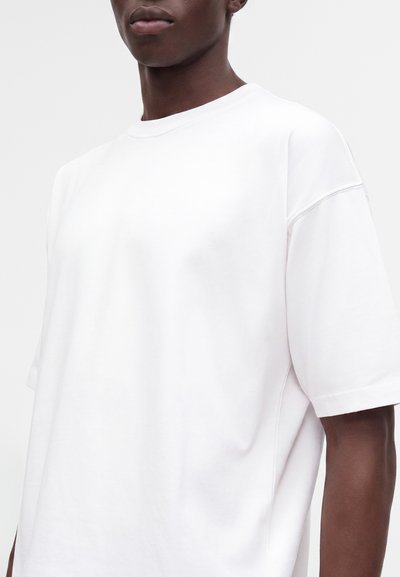 Personne à la peau foncée portant un t-shirt blanc uni oversize à manches courtes sur un fond gris clair.