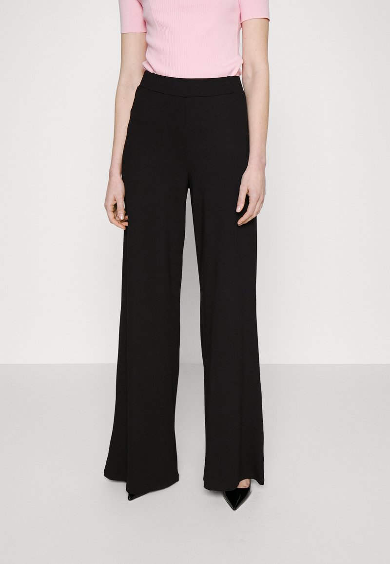 Femme portant un pantalon noir taille haute à jambes larges et un haut rose clair à manches courtes, debout sur un fond clair uni.