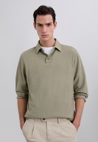 Camisa polo de manga larga en verde oliva claro con cuello y tapeta de tres botones, que cuenta con una tela texturizada y mangas enrolladas.