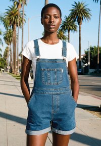 Junge Person steht auf einem sonnenbeschienenen Bürgersteig, der mit Palmen gesäumt ist, trägt ein weißes T-Shirt und Jeans-Latzshorts und hat die Hände in den Taschen.