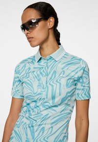 Jasnoniebieska koszulka polo z abstrakcyjnym, falistym wzorem w ciemniejszym odcieniu niebieskiego. Krótkie rękawy, kołnierzyk z białymi akcentami oraz rozporkiem na guziki.