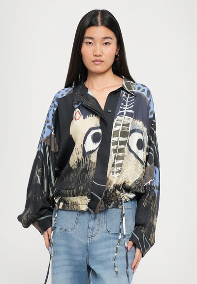 Woman with long dark hair wearing oversized shirt with abstract face design and light blue jeans, standing against white background.