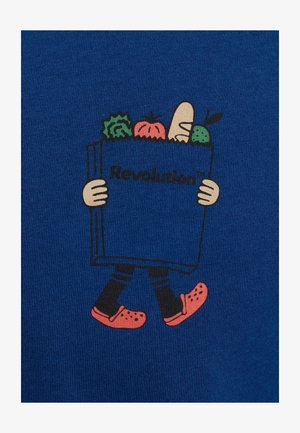 Blauwe stof met cartoonfiguur die een boek vasthoudt met de tekst "Revolutie!", bedekt met groenten en rode klompen draagt.