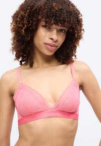 Νεαρή γυναίκα με σγουρά μαλλιά που φοράει ροζ μπραλέτα με δαντελωτό τελείωμα και λεπτές ρυθμιζόμενες τιράντες, σε απλό φόντο.