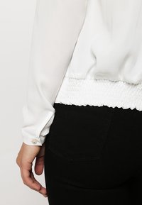 Weißes, leichtes Oberteil mit langen Ärmeln, gerafftem Taillendetail und knöpfbarem Manschetten. Kombiniert mit schwarzen, eng anliegenden Jeans aus glattem Material.