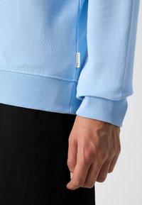 Light blue sweatshirt with ribbed cuffs and hem, featuring a subtle tag. Smooth texture and relaxed fit, paired with black fabric below.