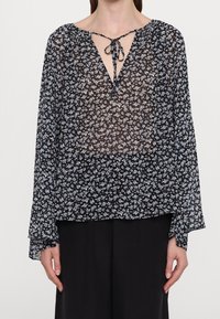 Femme portant un chemisier fleuri noir transparent à manches longues avec un lien noué à l'avant au niveau du col, associé à un pantalon noir.