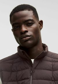 Gilet imbottito marrone scuro con colletto alto e chiusura zip. Presenta una texture trapuntata e tessuto liscio. Indossato sopra una camicia chiara.