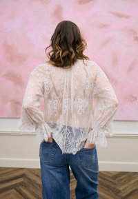 Femme aux cheveux ondulés portant un chemisier en dentelle blanche transparente et un jean bleu, debout les mains dans les poches arrière, devant un fond rose abstrait.