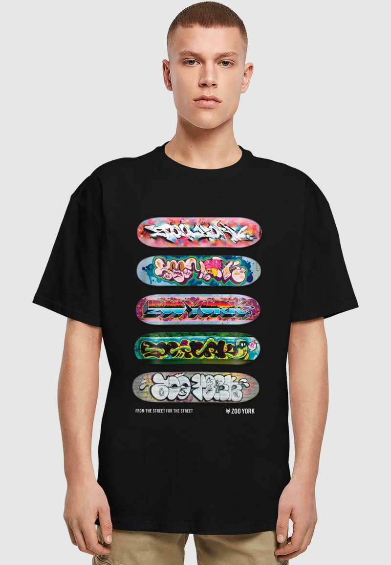 Camiseta negra con cinco patinetas coloridas que presentan texto y diseños al estilo graffiti, centradas sobre un fondo liso.