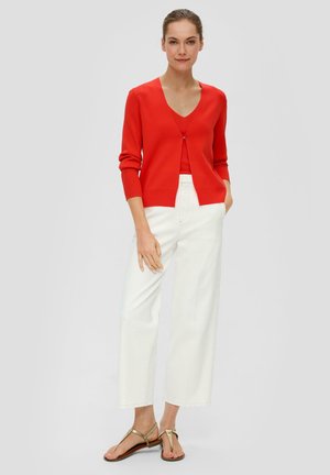 Cardigan en maille rouge avec un col en V, présentant un petit fermoir en argent. Associé à un pantalon large blanc et des sandales plates beiges.