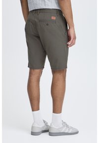 Olive green shorts made of lightweight fabric, featuring a back pocket with button detail and a leather patch label. Worn with gray sneakers.