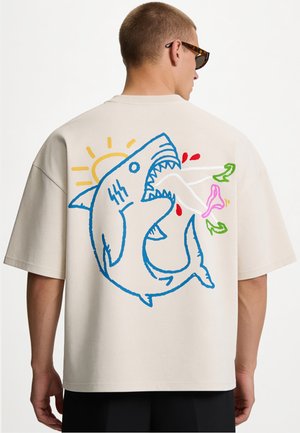 Personne portant un t-shirt beige oversize avec un requin cartoon coloré mordant un objet rose dans le dos, avec un détail soleil et gouttes rouges.