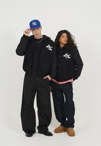 Zwarte jassen met wit logo, relaxte pasvorm, gecombineerd met zwarte broeken en bruine schoenen. Blauwe pet, casual stijl, met één model dat een roze shirt eronder draagt.
