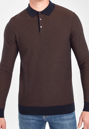 Mann trägt ein braunes, strukturiertes Langarm-Poloshirt mit navyfarbenem Kragen und Manschetten, kombiniert mit blauen Jeans, Torso sichtbar.