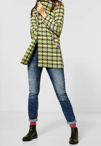 Cappotto a scacchi multicolore in giallo, verde e blu navy, abbinato a jeans in denim e stivaletti neri. Il cappotto presenta un colletto alto e bottoni.