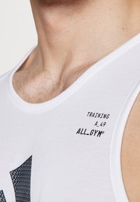 Bílý sportovní tílko s černým potiskem textu: "TRAINING A_49 ALL_GYM®." Materiál je hladký, s kulatým výstřihem a bezrukávovým provedením.