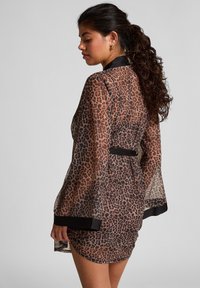 Vrouw met een krullende paardenstaart, gekleed in een doorschijnende jurk met luipaardprint en zwarte rand, poseert zijwaarts tegen een effen grijze achtergrond.