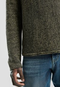 Gebreide trui in donkergroen en zwart met een gestructureerd patroon, voorzien van een ronde halslijn en lange mouwen, gecombineerd met blauwe denim jeans.