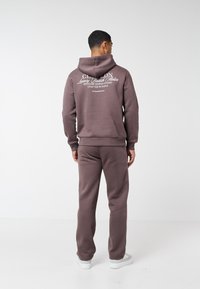 Egy férfi háttal áll, lila kapucnis pulóvert és hozzáillő melegítőnadrágot visel, a kapucnis pulóver hátán fehér felirat látható, fehér padlón áll, egyszerű háttér előtt.