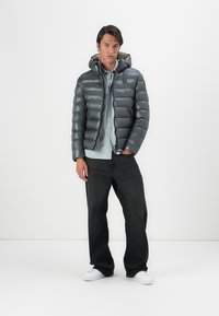 Gray puffer jacket with a hood, featuring a shiny, quilted design. Worn over a light blue shirt and paired with dark jeans and white shoes.