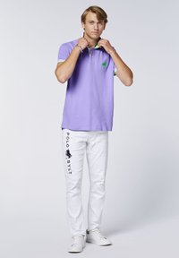 Helles lila Polohemd mit grünen Akzenten, kurzen Ärmeln und Logo. Weiße Hose mit dem Schriftzug "POLO SYLT" und einem dunklen Logodetail. Weiße Schuhe.