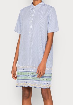 Mujer que lleva un vestido de manga corta, con cuello y botones, con rayas verticales azules y patrones bordados en verde y amarillo cerca del dobladillo.