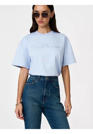 Naine kannab helesinist lühikeste varrukatega särki kirjaga „See Moves“ ja kõrge vöökohaga siniseid teksaseid, aksessuaaridena päikeseprille ja käepaela.