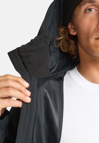 Schwarze Jacke mit einem strukturierten Innenfutter der Kapuze, glatter Außenseite und Netz-Innenfutter. Ein weißes T-Shirt ist darunter sichtbar.