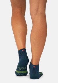 Calcetines tobilleros azul marino con detalles verdes, que cuentan con un puño acanalado y suelas texturizadas, mostrados en las piernas de una persona de pie.
