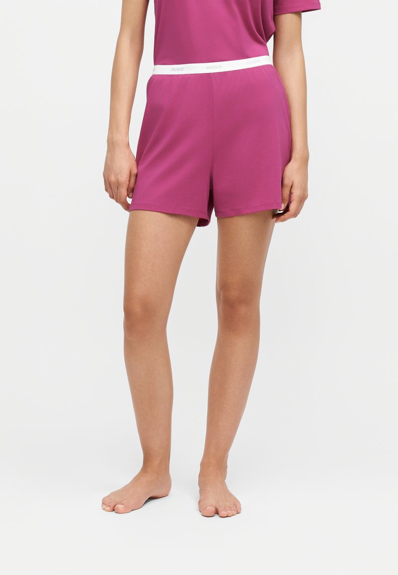 Personne portant un short rose et un haut rose assorti, debout pieds nus sur un fond blanc uni.