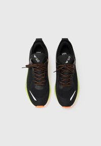 Pair of black athletic sneakers with orange and black laces, green soles, white details, and brand logos on the tongue, displayed top-down.