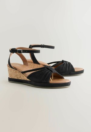 Czarne sandały na koturnie z zamszowej górą, korkowym obcasem i wiązanym przodem. Posiadają regulowany pasek wokół kostki oraz teksturowaną wkładkę dla większego komfortu.
