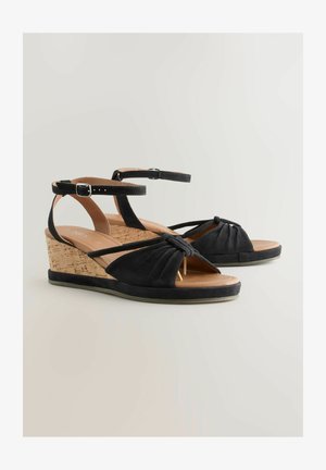 Melnas platformas sandales ar zamšādas augšdaļu, korķa papēdi un mezgla priekšējo dizainu. Aprīkotas ar regulējamu potītes siksniņu un strukturētu kājstarpi komfortam.