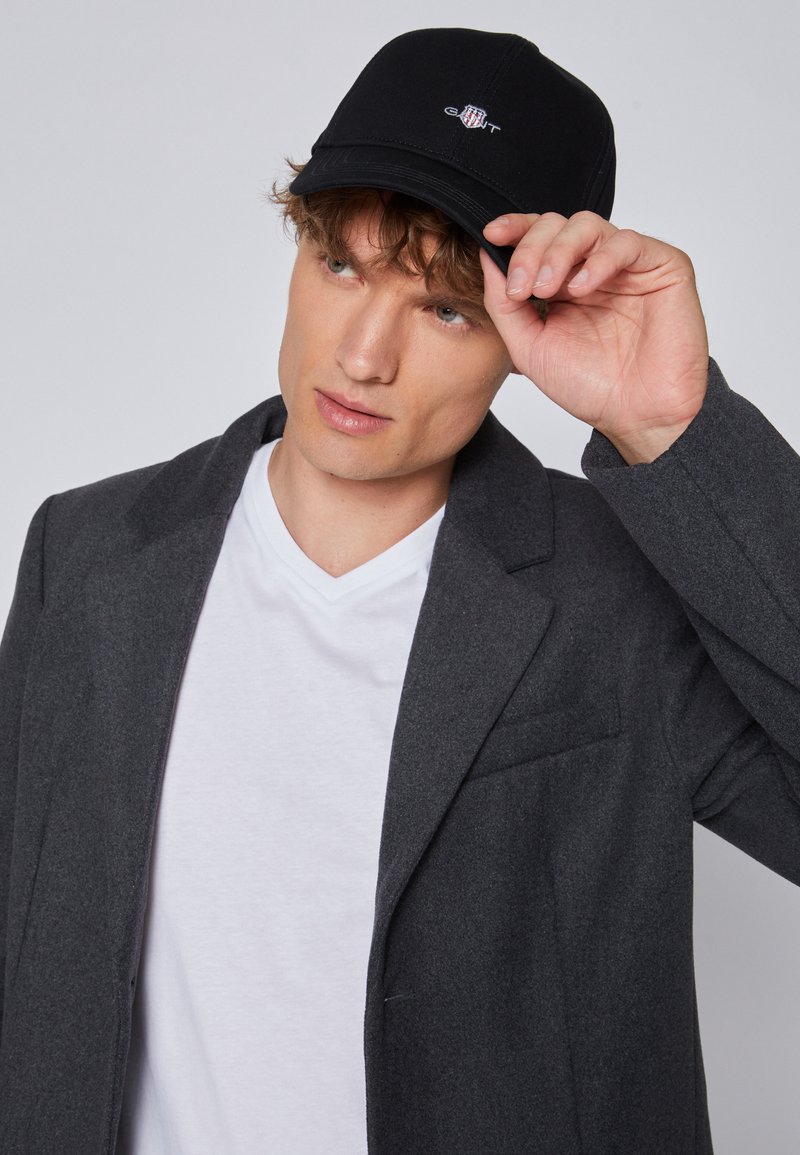 Gray wool blend coat with notch lapels, worn over a white V-neck t-shirt. A black cap with a logo is held on the head. Short, wavy hair visible.