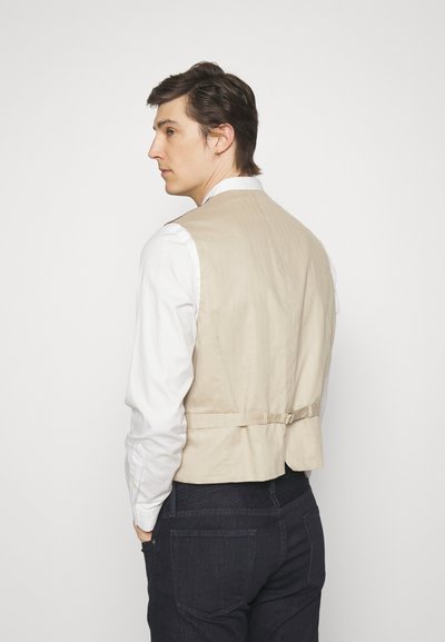 Beżowa, dopasowana kamizelka wykonana z lekkiego materiału, z detalem z tyłu w postaci klamry, noszona na białej koszuli i ciemnych dżinsach.