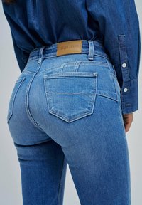 Jeans de mezclilla azul con una etiqueta de cuero en la cinturilla, dos bolsillos traseros y un diseño ajustado con un acabado ligeramente desgastado.