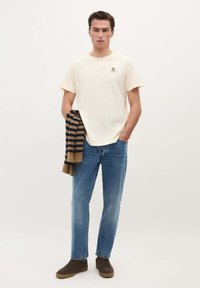 Beige bomulds T-shirt med korte ærmer og et lille kranium logo. Båret sammen med blå lige ben jeans og en stribet brun og marineblå sweater draperet over den ene arm.