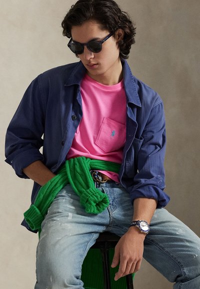 Μπλε πουκάμισο με κουμπιά, ροζ μπλουζάκι με τσέπη, πράσινο πλεκτό πουλόβερ δεμένο γύρω από τη μέση, ανοιχτό μπλε τζιν τσαλακωμένα, ρολόι στον καρπό, γυαλιά ηλίου.